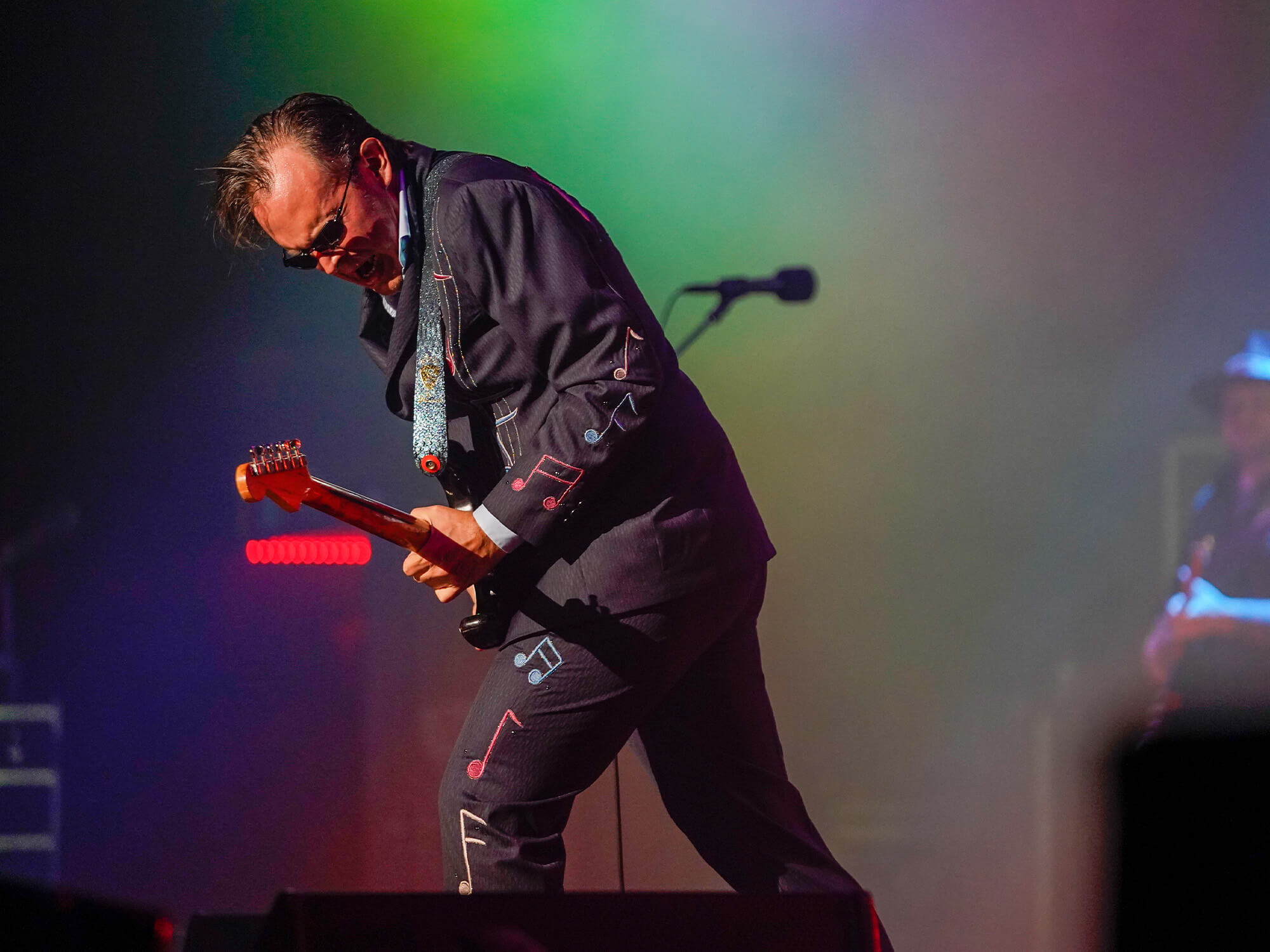 Joe Bonamassa on stage playing guitar. He is slightly hunched over his instrument, is wearing his signature sunglasses and suit, and is playing with a passionate expression on his face.